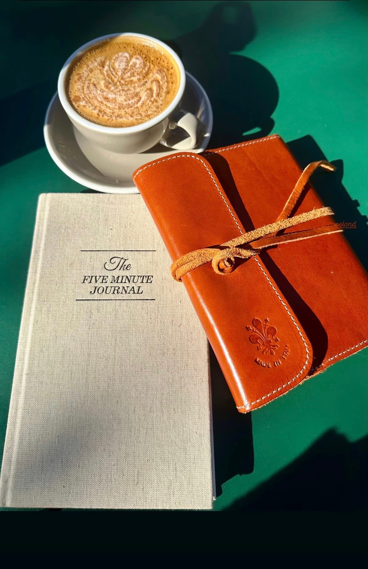 A latte with leaf-pattern foam art beside The Five Minute Journal and a leather journal tied with cord, on a green tabletop in sunlight